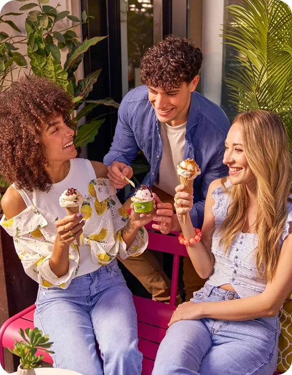 Three friends on a patio laugh together while holding ice cream cones and a cup, surrounded by lush green plants.