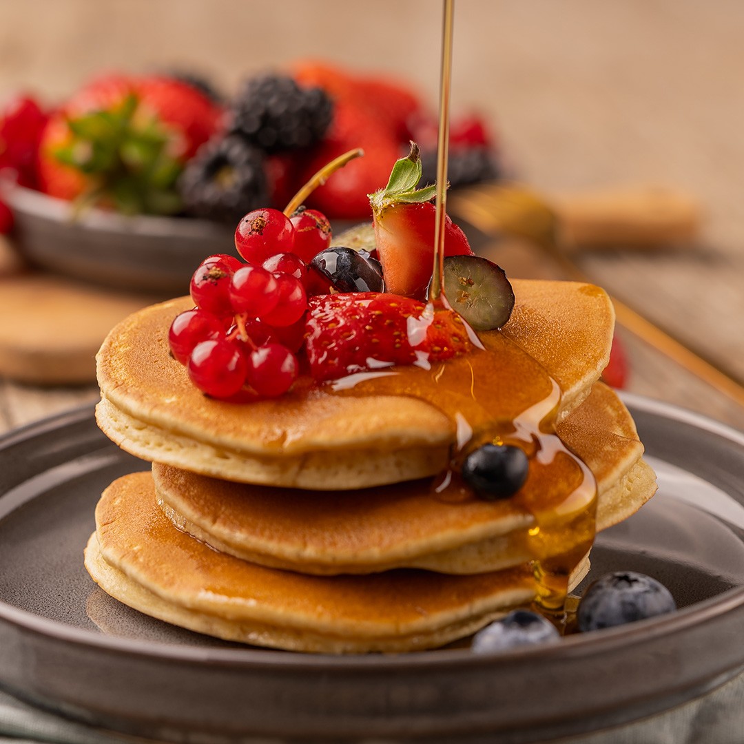 Sirope dorado cayendo sobre un stack de panqueques con fresas, arándanos, moras y grosellas rojas