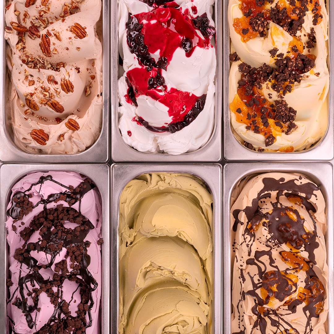 Seis bandejas metálicas de gelato en diferentes sabores, incluyendo pecana, frutos rojos, pistacho y chocolate con ca...